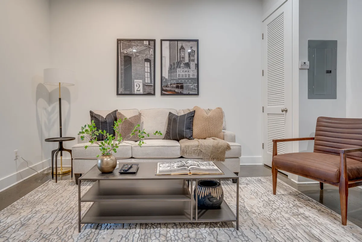An interior shot of a cozy lounge area featuring a comfortable sofa with decorative pillows, a coffee table with a book, wall artwork, and a stylish floor lamp. The atmosphere is contemporary and homey, perfect for relaxing or socializing.