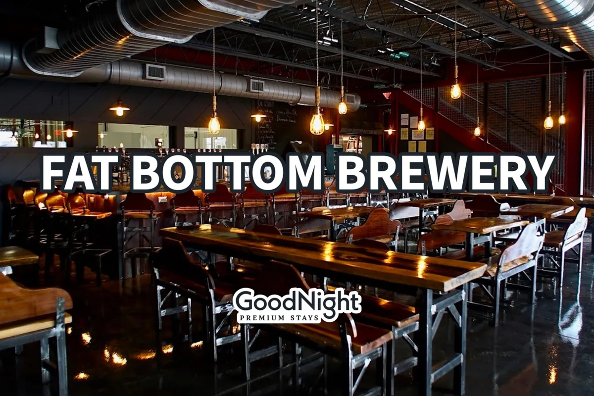 The interior of a modern brewery features a long wooden bar with stools, warm suspension lighting, and brewery equipment in the background. Groups of people are seen socializing, tasting craft beers, and enjoying casual dining in a lively, relaxed setting.