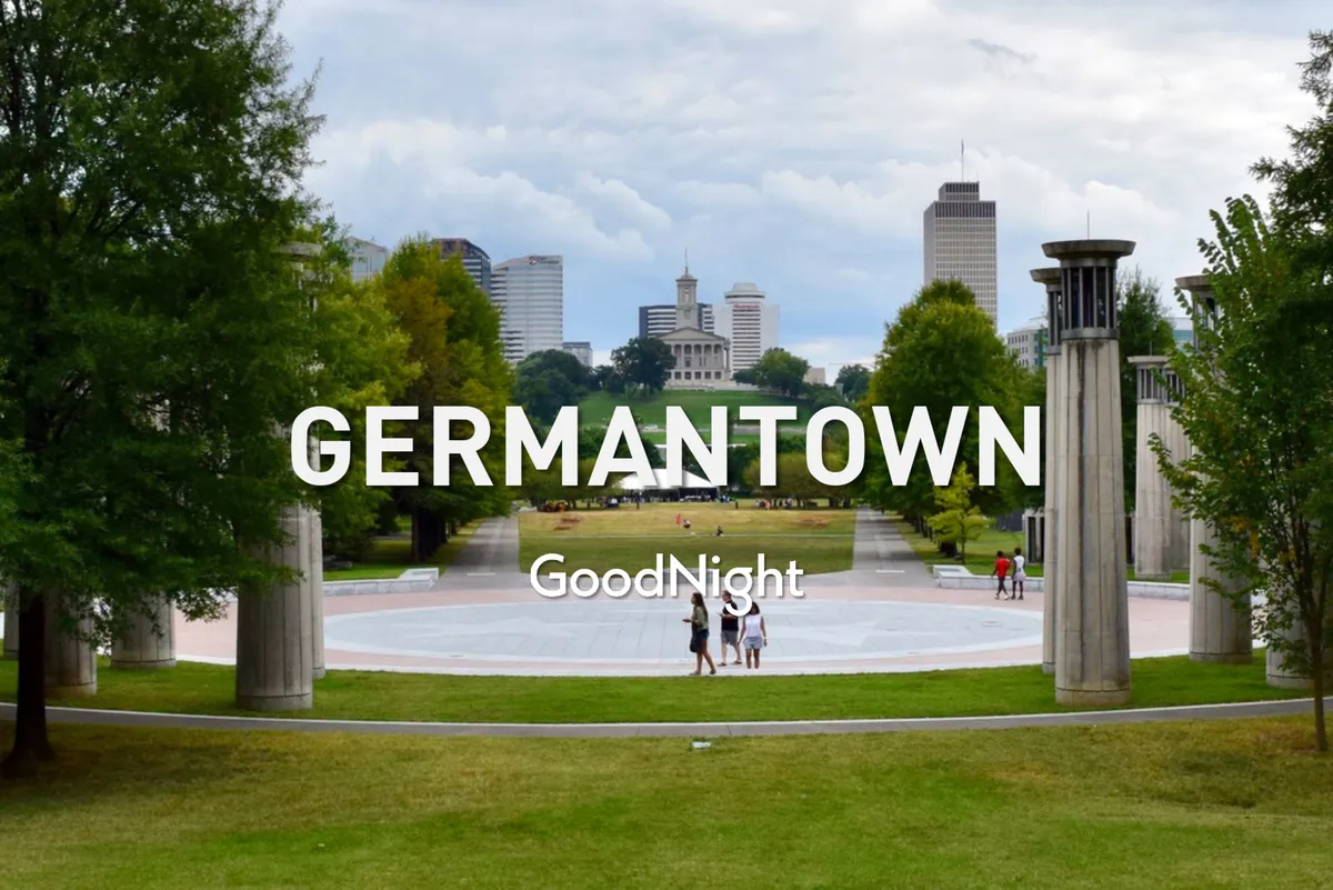 A wide, open green space in a city park with the Nashville skyline in the distance and classical archway columns, perfect for an evening stroll or picnic. The area is bathed in soft evening light, inviting couples and small groups to relax and enjoy the tranquil atmosphere.