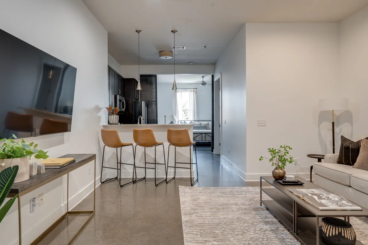 This image features a contemporary living area with a television mounted on the wall, bar stools lined up at the kitchen island, a stylish sofa decorated with pillows, and a coffee table holding magazines. The room is bathed in natural light, thanks to the large windows, and there's a clear visual connection between the lounge and kitchen, reinforcing the open-plan layout.
