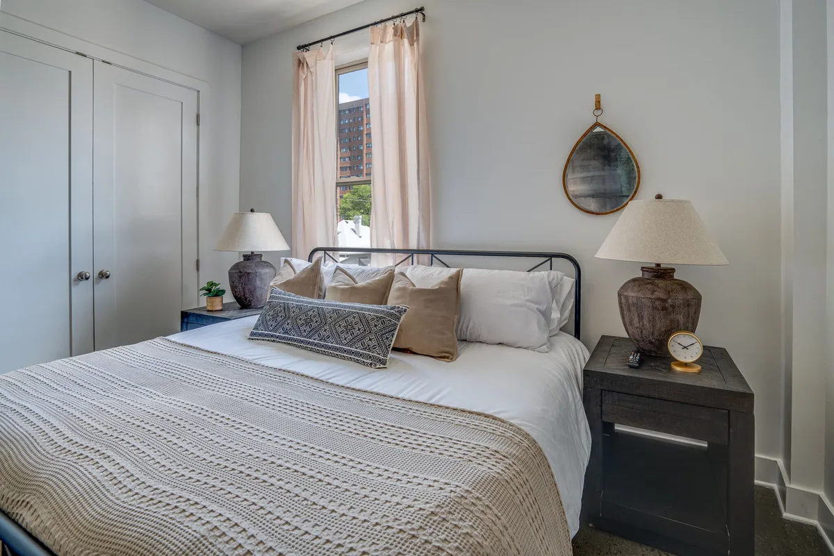 A tranquil bedroom showcasing a bed adorned with decorative pillows, nightstands on either side with warm lamps, and a window draped in sheer curtains letting in natural light. The overall look is inviting and peaceful, emphasizing comfort and relaxation.